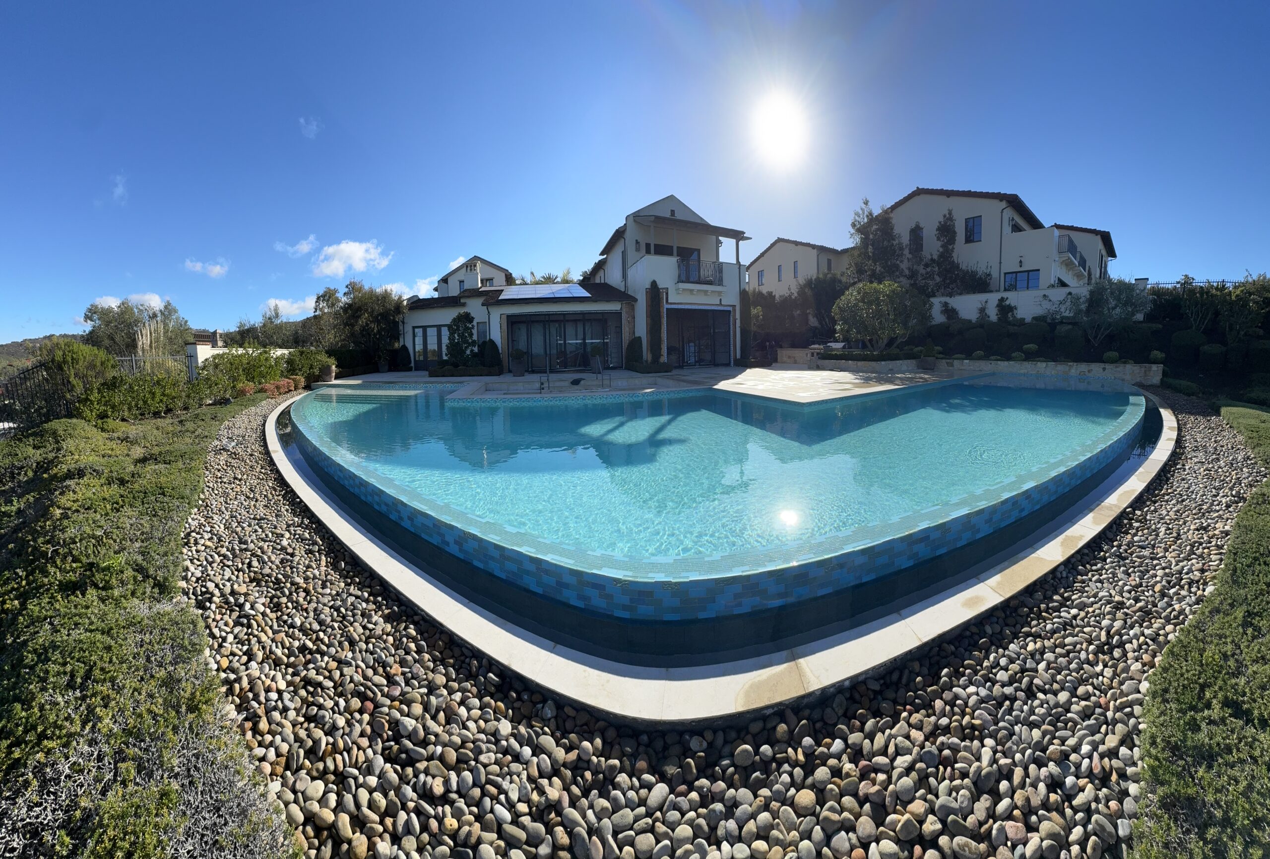 IMG_9909 Large outdoor swimming pool with clear water, surrounded by rocks and greenery, set in front of Mediterranean-style houses under a bright sunny sky.